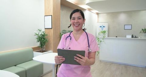 Smiling nurse holding tablet in modern clinic reception wearing pink scrubs and stethoscope