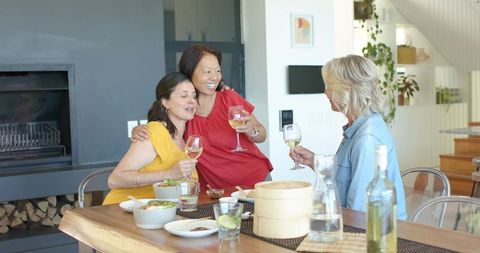 Senior asian woman celebrating with two female friends clinking wine glasses at home dining table