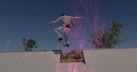 Skateboarder performing airborne trick over concrete gap with pink powder burst, blue sky