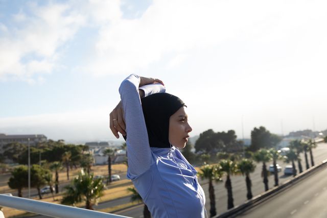 Biracial woman wearing hijab and sportswear stretching outdoors on a sunny day. She is listening to music with earphones while exercising in an urban environment. Ideal for use in content related to fitness, healthy living, urban lifestyle, and diversity in sports.