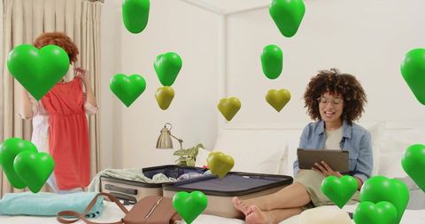 Woman using tablet while packing suitcase, friend holding dress, green heart overlays
