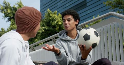 Two young men chatting on campus steps while holding soccer ball, casual university lifestyle