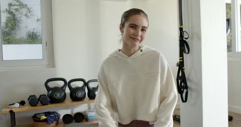 Smiling woman in home gym wearing cream hoodie and maroon pants with kettlebells
