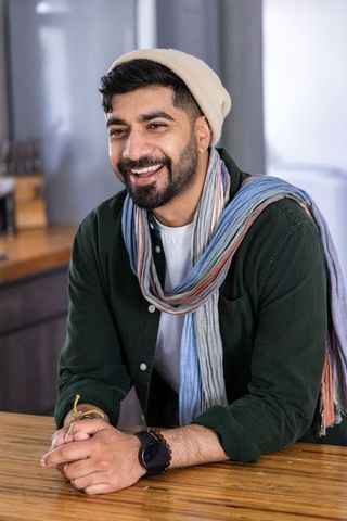 Indian man smiling while leaning on wooden kitchen counter wearing beanie and scarf