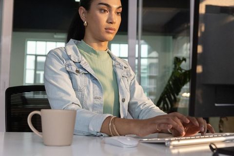 Focused professional woman typing at modern office desk with coffee mug, glass partitions