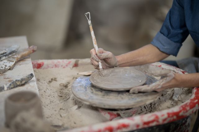 Close-up of a female potter using a hand tool to mold a clay plate on a pottery wheel. Her hands are covered in clay, showcasing the detailed craftsmanship involved in pottery making. This image is perfect for use in articles or advertisements related to arts and crafts, pottery workshops, handmade ceramics, and traditional craftsmanship.