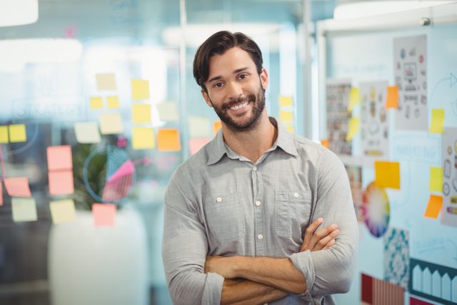 Young male executive standing confidently with arms crossed in a modern, creative office. Ideal for business websites, office culture articles, modern work environment concepts, and leadership promotions.