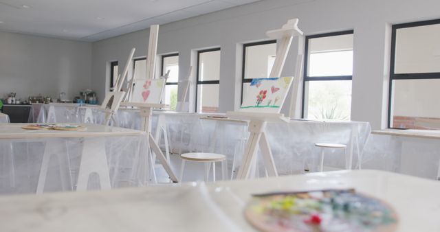 Featuring a serene, well-lit art classroom with easels set up with unfinished paintings. Ideal for use in educational materials, advertisements for art classes, or inspirational creative content. Evokes a sense of creativity and anticipation in an empty studio setting.