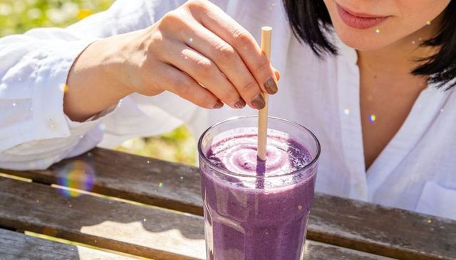 Woman in white shirt stirring purple berry smoothie with bamboo straw on wooden table