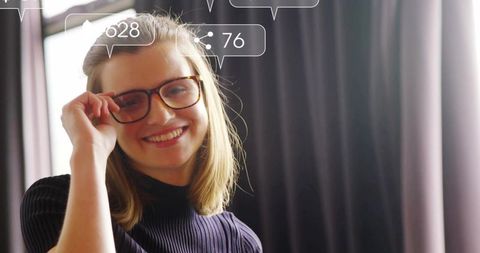 Smiling woman adjusting tortoise-frame glasses by window with social media icon overlays