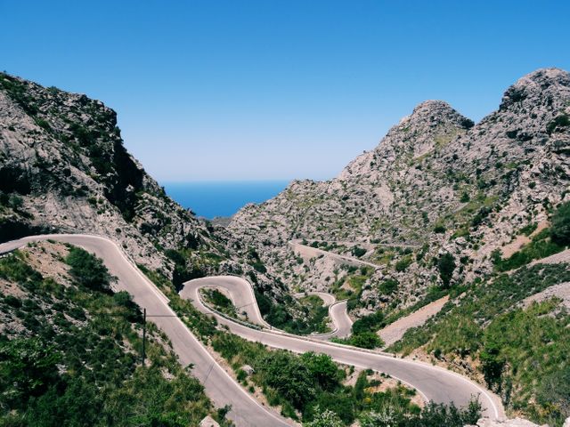 Winding mountain road leading towards the ocean under clear blue sky. Ideal for travel blogs, tourism websites, adventure promotions, and landscape wallpapers. Highlights adventurous road trips and scenic drives with stunning natural views.