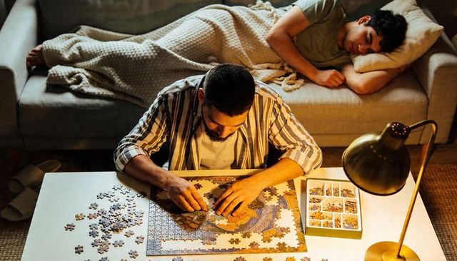 Man assembling jigsaw puzzle on living room table with sleeping friend nearby cozy lamp