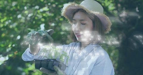 Woman wearing straw hat inspecting seedling with digital data overlay for smart gardening