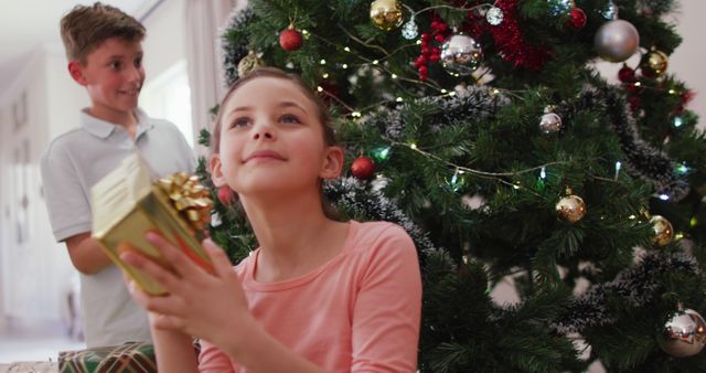 Children are unwrapping gifts by a beautifully decorated Christmas tree, creating a joyful and exciting holiday moment. Useful for festive promotion, holiday greeting cards, family celebration themes.