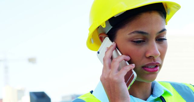 A young African American woman in a hard hat is focused on a phone conversation, with copy space. Her profession appears to be in the construction or engineering field, indicated by her safety gear.