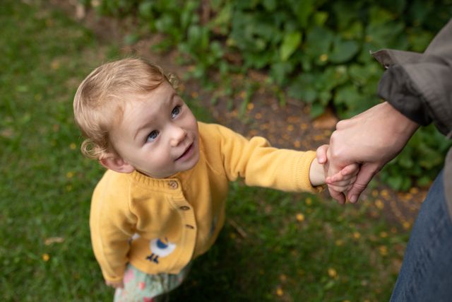 High angle view of a Caucasian baby standing in a garden, holding her mums hand, looking at her and smiling.