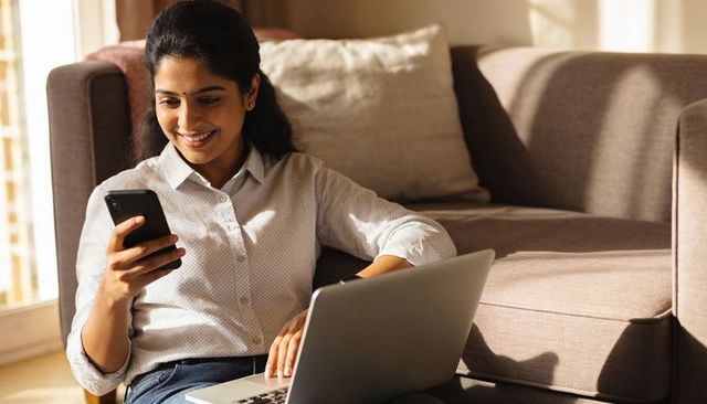 Sunlit indian woman working from home with laptop and smartphone on cozy living room floor