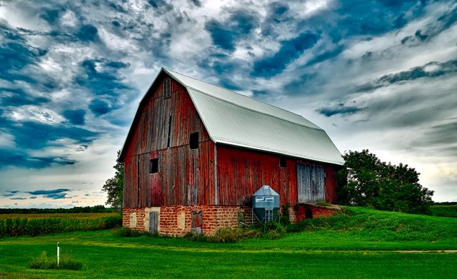 This image shows an old wooden barn standing prominently on a lush green field with a dramatic, cloudy sky overhead. The barn has a classic vintage look with red weathered planks and a metal roof. Perfect for use in projects related to farming, rural life, nostalgia, traditional architecture, and agriculture. Ideal for ads, blogs, websites, or magazines focusing on countryside scenics, heritage preservation, and rustic themes.