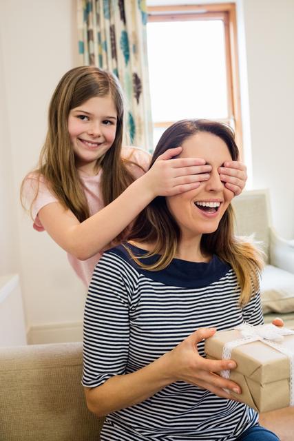 Daughter covering her mother's eyes while giving a surprise gift in a cozy living room at home. Ideal for use in family-oriented advertisements, parenting blogs, Mother's Day promotions, and articles about family bonding and celebrations.