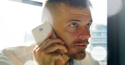 Mid-adult man talking on smartphone at office window with sunlit bokeh and lens flare