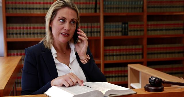 Female lawyer consulting on the phone while referencing a law book in an office library. The setting is professional, focusing on the legal environment with law books and a gavel visible. Ideal for articles on legal advice, professional consulting services, law firm promotions, or legal industry websites.