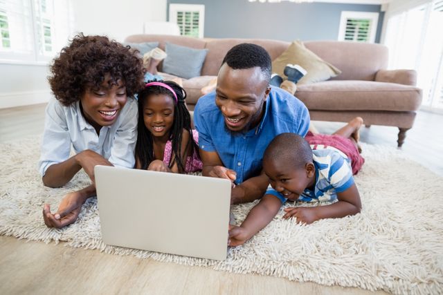 African American family of four lying on carpet in living room, smiling and using laptop together. Ideal for concepts related to family bonding, technology use at home, leisure activities, and modern family life.