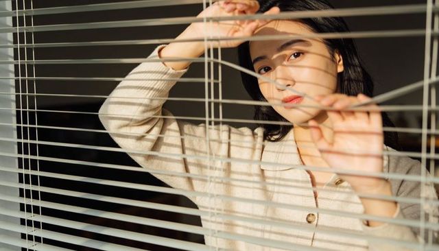 Asian woman peering through blinds in warm sunlight, striped shadows, thoughtful expression