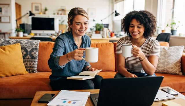 Two women collaborating and reviewing charts on laptop while drinking coffee on orange sofa