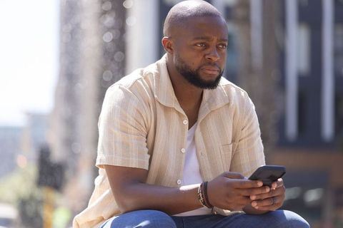 Contemplative man sitting in sunlit urban plaza holding smartphone, casual summer outfit