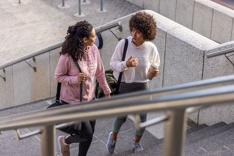 Two friends walking and talking up urban stairs after workout carrying duffel bags and coffee