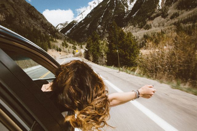 Woman with wavy hair enjoying a road trip by leaning out car window and extending arm on a scenic mountain highway. Ideal for depicting travel, adventure, and freedom. Suitable for articles, brochures, and advertisements related to travel and tourism, car rentals, and outdoor activities.
