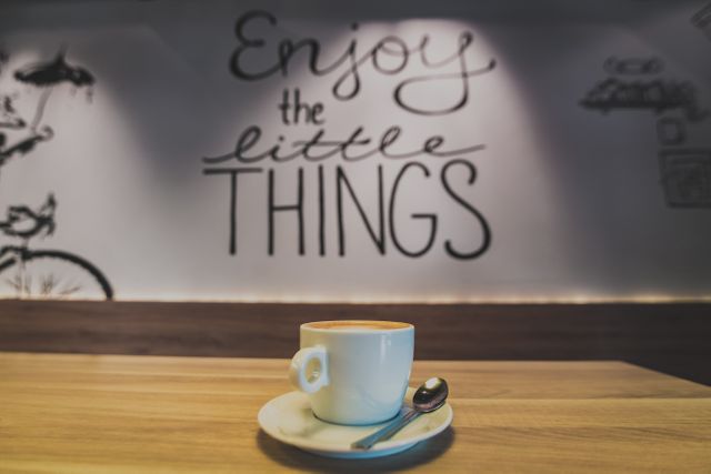 Coffee cup with saucer and spoon on wooden table in cozy cafe. Background features motivational wall art with 'Enjoy the little things' message in elegant handwriting. Ideal for promoting cafes, relaxation, positivity, or the importance of small pleasures in life.