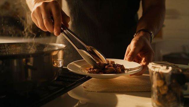 Man plating hearty stew with tongs on white plate beside steaming pot in cozy kitchen