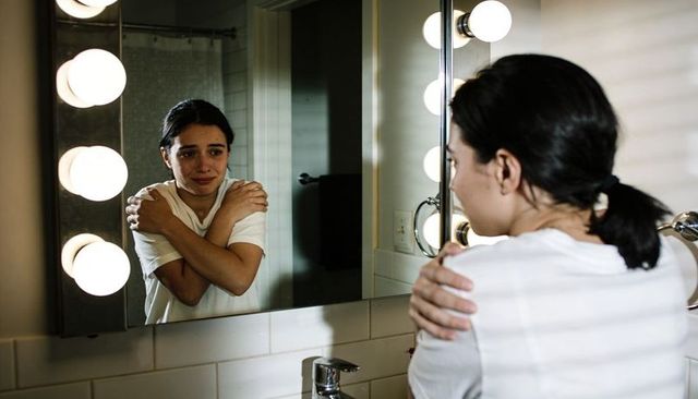 Young woman comforting herself hugging shoulders in bathroom mirror with vanity lights
