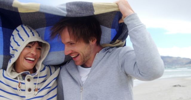 A young Caucasian couple enjoys a playful moment under a blanket at the beach, with copy space. Their laughter and closeness suggest a romantic and carefree connection amidst a breezy seaside setting.
