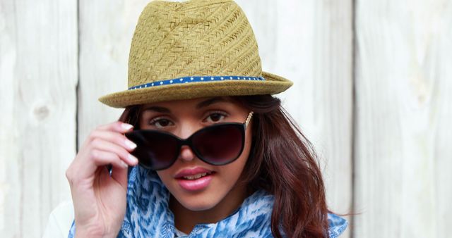 Young woman wearing a straw hat and sunglasses, touching the eyewear with one hand, posing against a rustic wooden background. She is also accessorizing with a scarf, creating a casual and summery look. This image can be used for fashion blogs, advertisements for summer accessories, or social media campaigns focused on casual chic dressing.