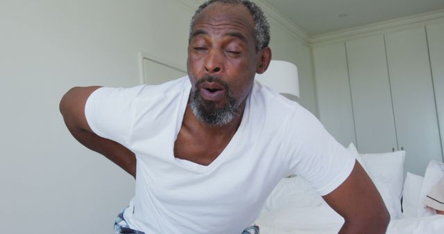 Elderly man showing discomfort by holding his lower back in a bright bedroom. Useful for content related to aging, health issues, back pain, chiropractic care, senior health, home medical solutions, and lifestyle of mature adults.
