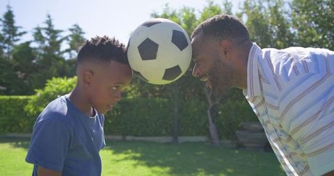 African american father and son balancing soccer ball between foreheads in sunny backyard