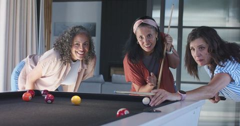 Three women playing pool on black-felt table in modern lounge, friends laughing