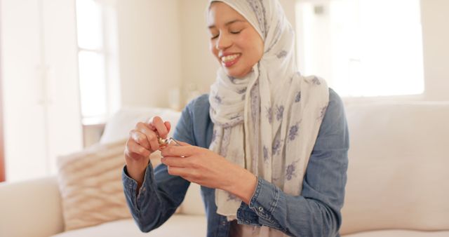 Happy biracial woman in hijab smelling perfume at home, copy space. Lifestyle, domestic life, wellbeing concept, unaltered.
