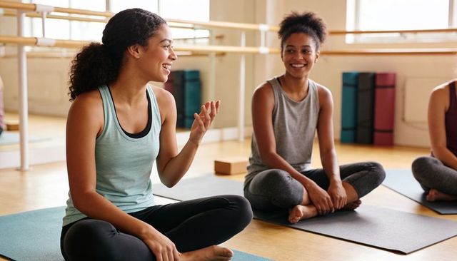 Young women chatting on yoga mats during post-class stretch in bright fitness studio