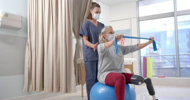 Senior woman with prosthetic leg receives assistance from physiotherapist while exercising with fitness band on a fitness ball. Great for topics on physical therapy, rehabilitation, senior healthcare, prosthetics, and fitness recovery.