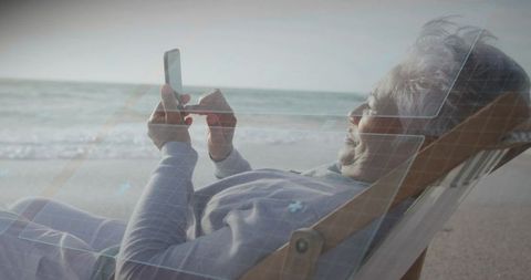 Senior woman reclining on beach using smartphone with digital overlay for remote lifestyle
