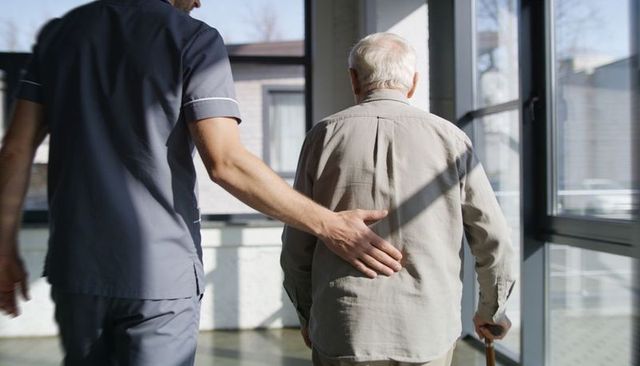 Caregiver assisting elderly man walking with cane in sunlit healthcare corridor