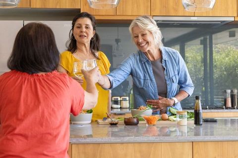 Diverse mature women cooking and toasting wine at kitchen island, friends gathering