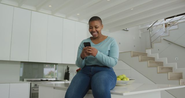 Happy african american plus size woman sitting on countertop in kitchen, using smartphone. domestic lifestyle, enjoying leisure time at home with technology.