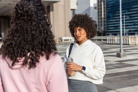 Female friends chatting on city plaza wearing hoodies holding water bottle and smartwatch