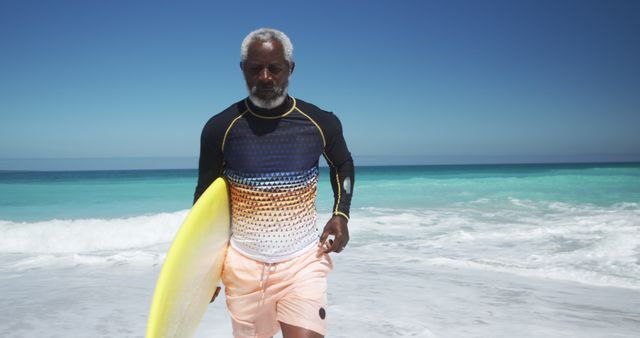 Elderly man standing on shore with surfboard while calm ocean waves lap at his feet. Perfect for content promoting active lifestyles, retirement activities, or ocean sports. Suitable for travel blogs, health and wellness, or advertisements focusing on senior fitness.