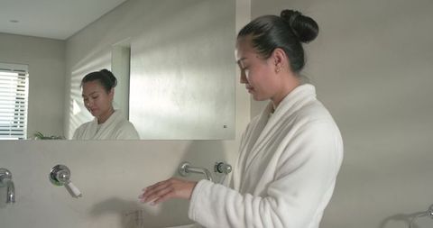 Mid-adult asian woman wearing white bathrobe using sink and mirror in minimalist bathroom