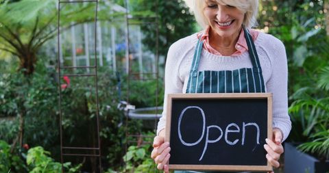 Happy Mature Gardener Holding 'Open' Sign in Lush Greenhouse - Download Free Stock Images Pikwizard.com